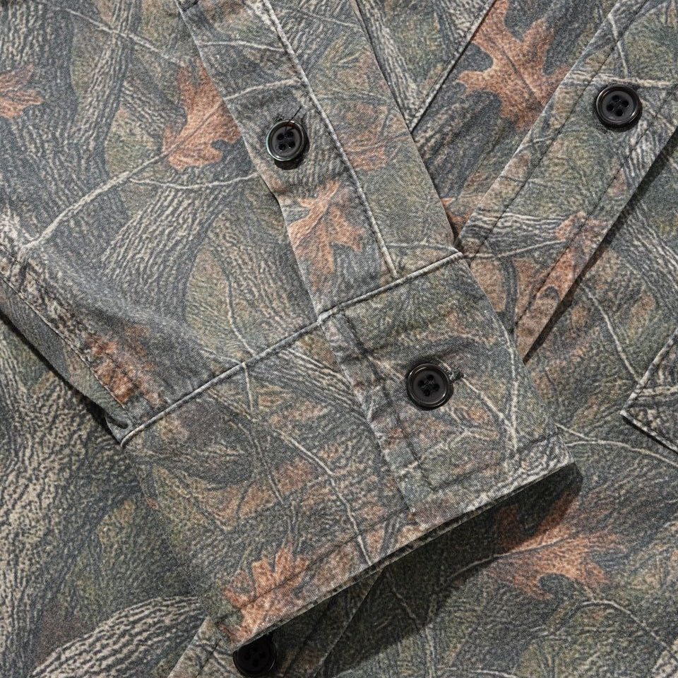 Close-up of a camouflage button-up shirt with a woodland leaf and bark pattern in muted green, brown, and gray tones, featuring dark buttons.