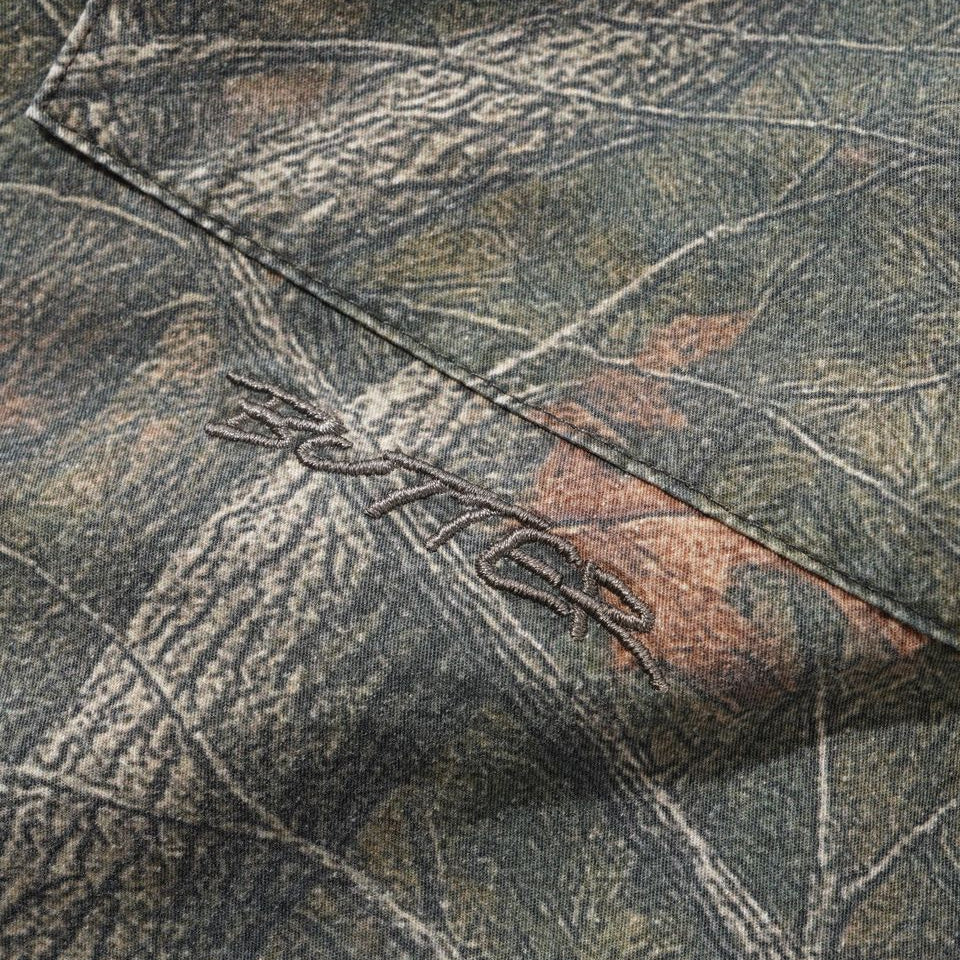 Close-up of a camouflage fabric with a tree bark and leaf pattern in muted green, brown, and gray tones, featuring a stitched logo that appears to read 'BUTTER'.