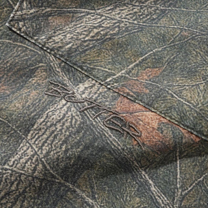 Close-up of a camouflage fabric with a tree bark and leaf pattern in muted green, brown, and gray tones, featuring a stitched logo that appears to read 'BUTTER'.