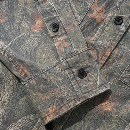 Close-up of a camouflage button-up shirt with a woodland leaf and bark pattern in muted green, brown, and gray tones, featuring dark buttons.