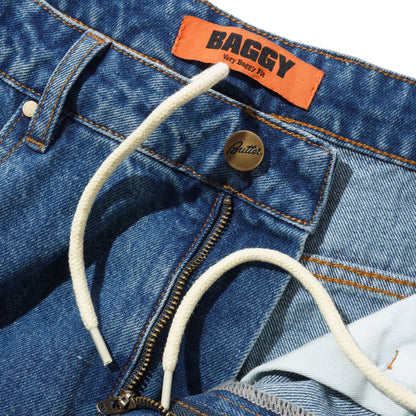 Close-up of a pair of blue denim jeans showing the waistband, orange BAGGY brand label, white drawstring, metal zipper, and a gold-tone button.