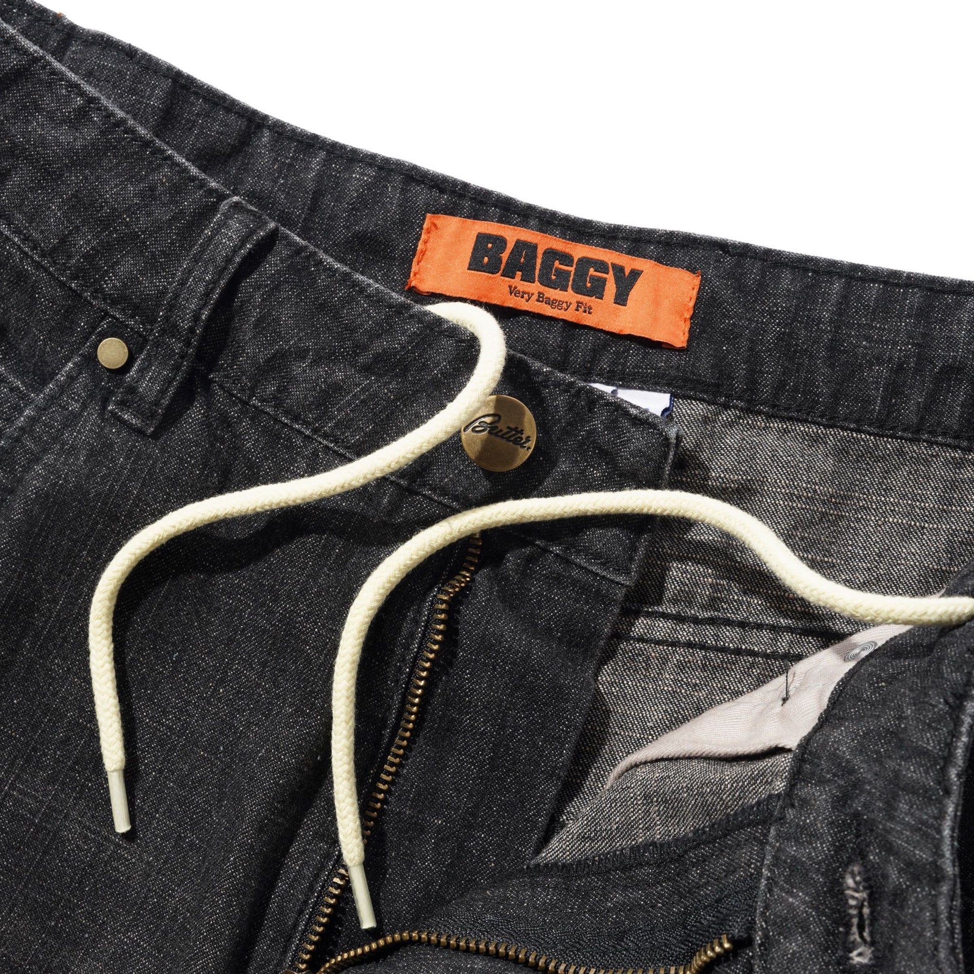 Close-up of dark gray denim drawstring pants with an orange BAGGY brand label, cream drawstring, and brass zipper on a white background.