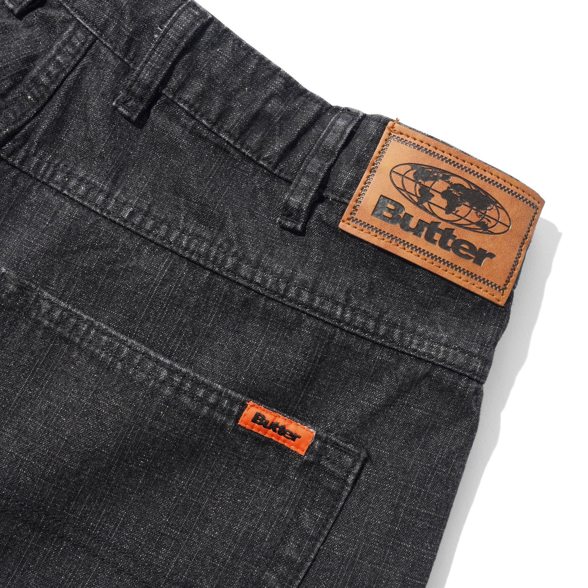 Close-up of dark gray denim jeans showing the back waistband with a large tan leather Butter patch and a smaller orange leather patch on the pocket.