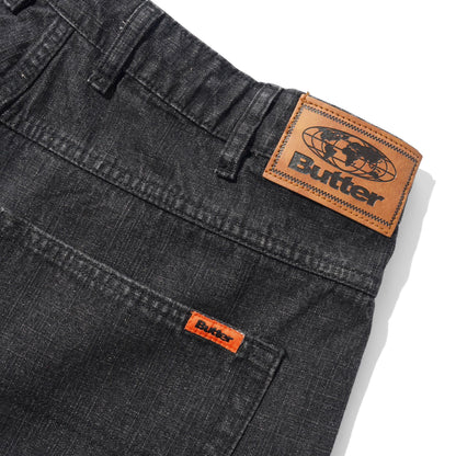 Close-up of dark gray denim jeans showing the back waistband with a large tan leather Butter patch and a smaller orange leather patch on the pocket.