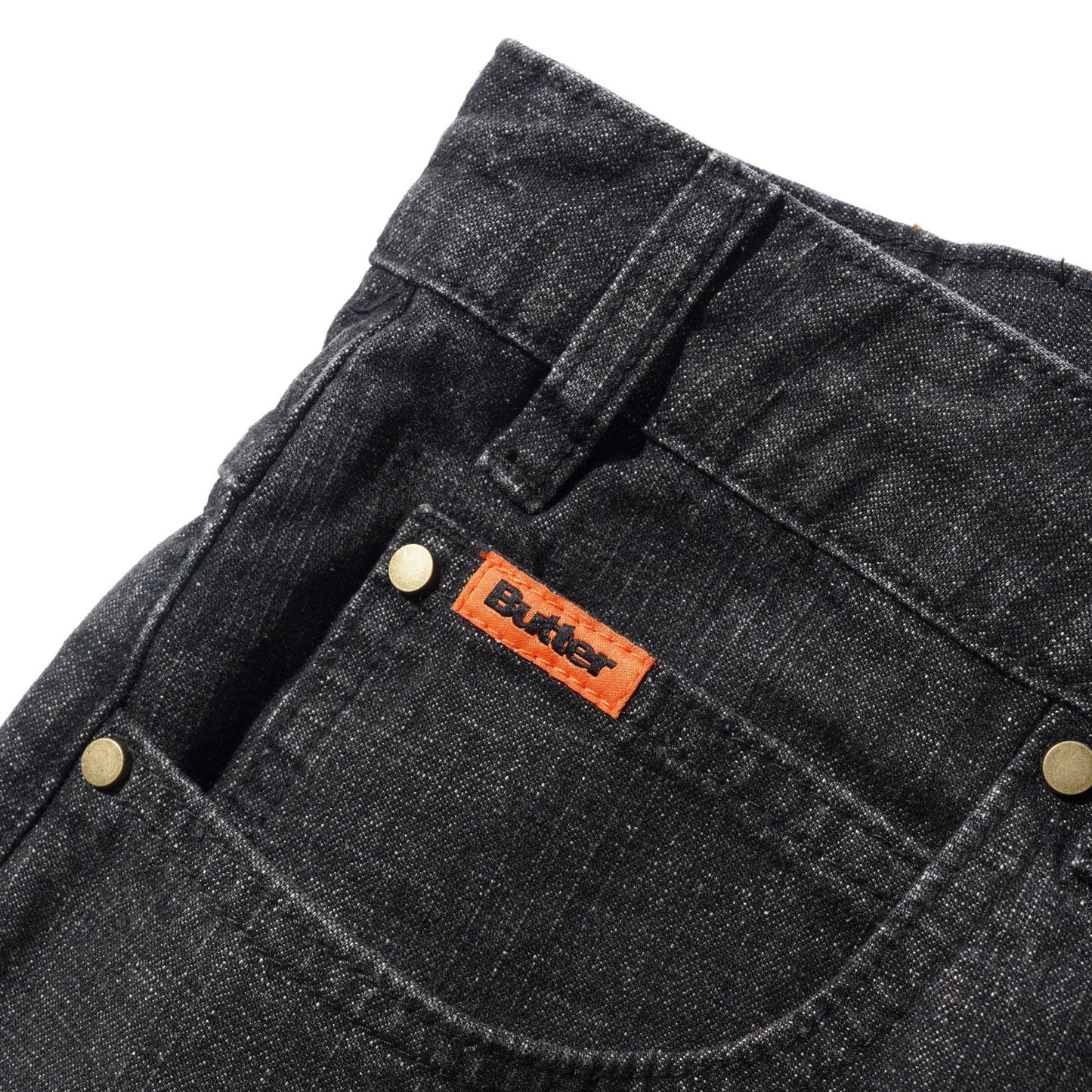 Close-up of the front pocket of dark gray washed denim jeans, featuring brass rivets and a prominent orange leather Butter brand patch.