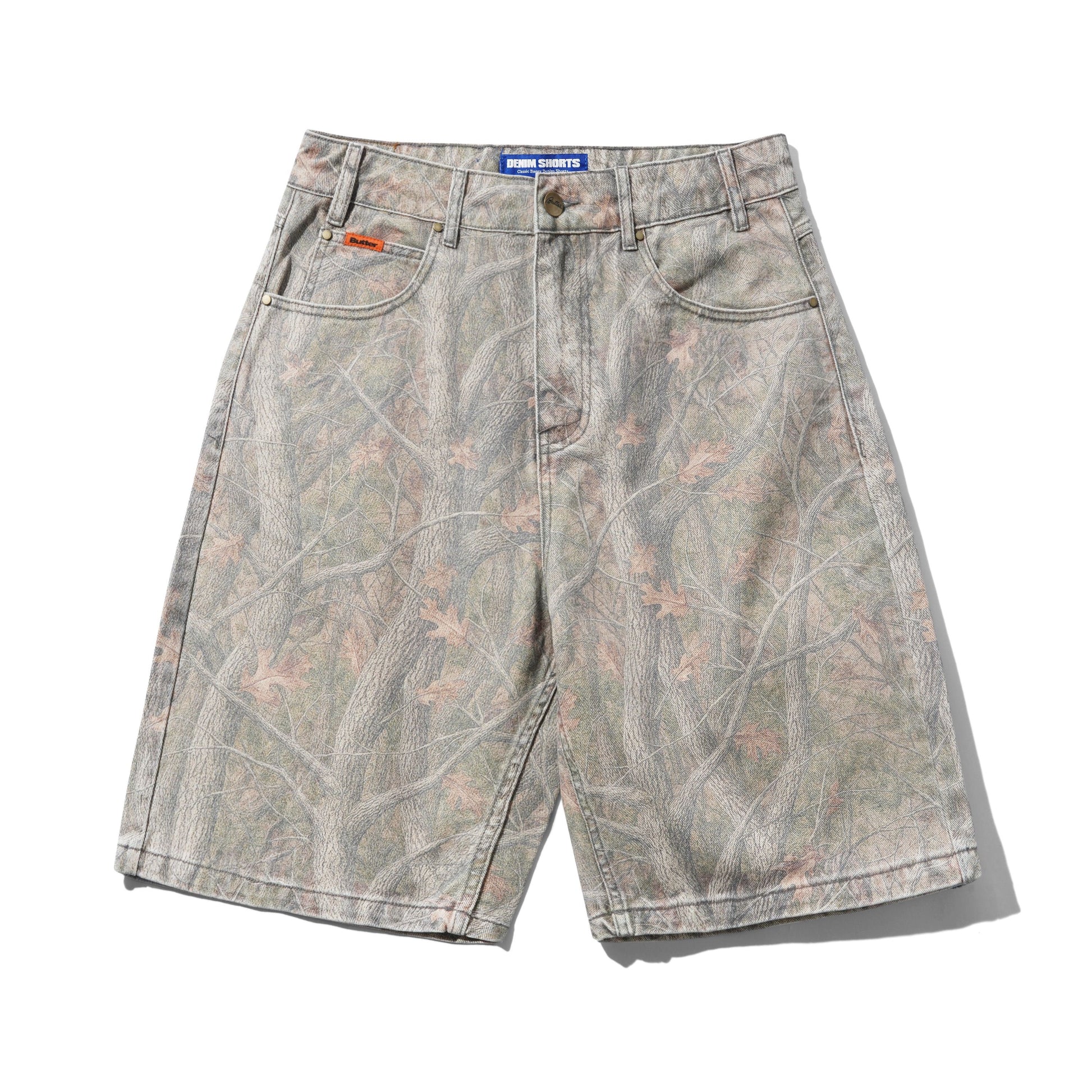 A pair of vintage-style denim shorts featuring a light gray and beige tree-bark camouflage pattern with subtle pinkish leaf details. The shorts have a faded wash, a button fly, belt loops, and a blue waistband label