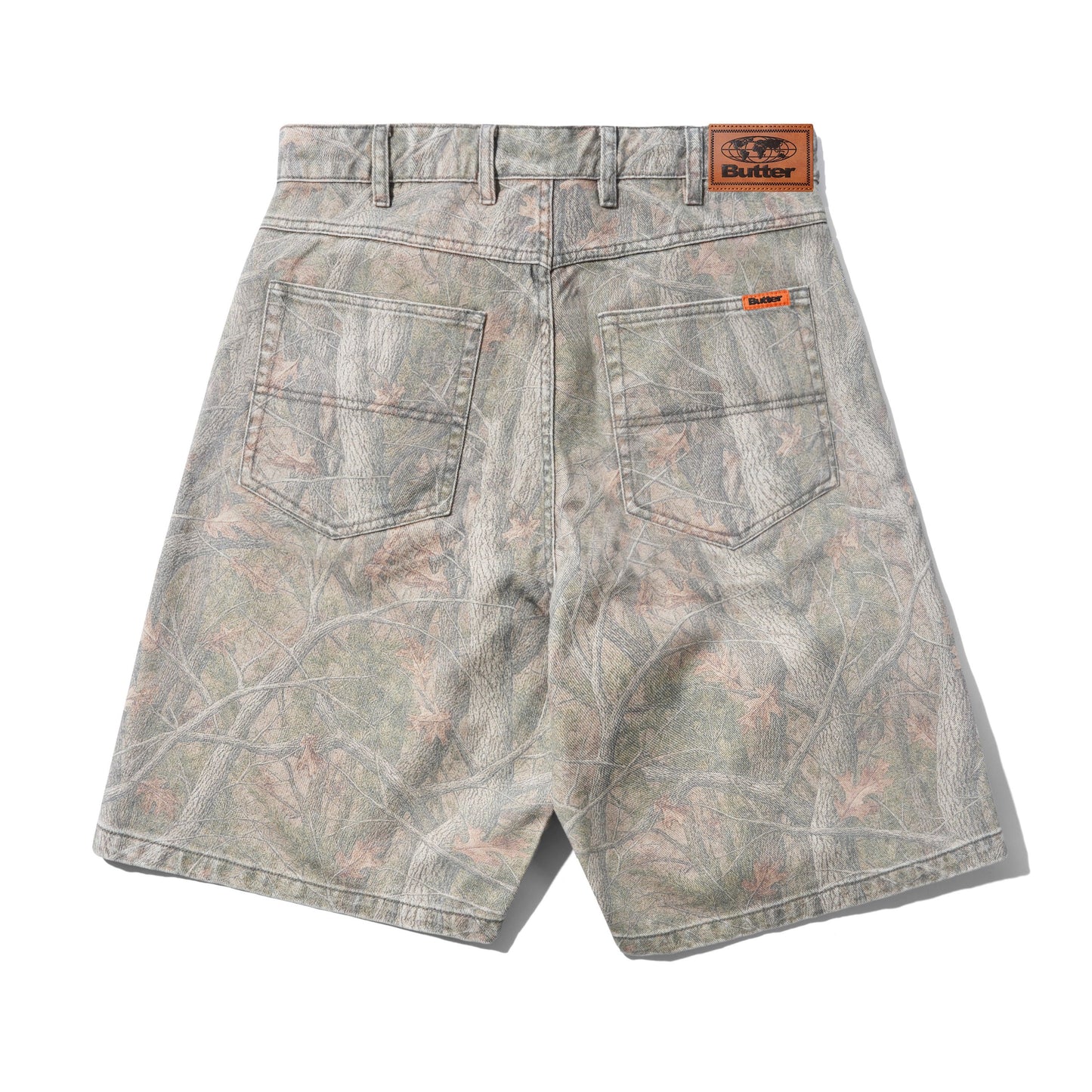 Back view of camouflage denim shorts with a gray, green, and brown woodland branch pattern, featuring a brown leather 'BUTTER' brand patch on the waistband and an orange brand tag on the back pocket.