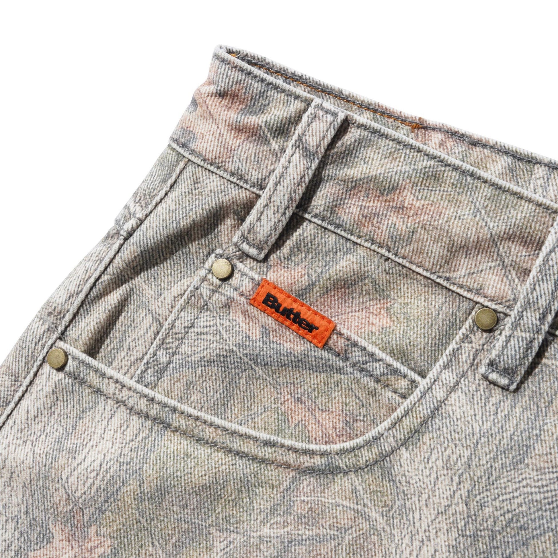 Close-up of the back pocket of a pair of faded gray denim jeans with a subtle pink and beige floral pattern, featuring a signature orange Butter brand tab.