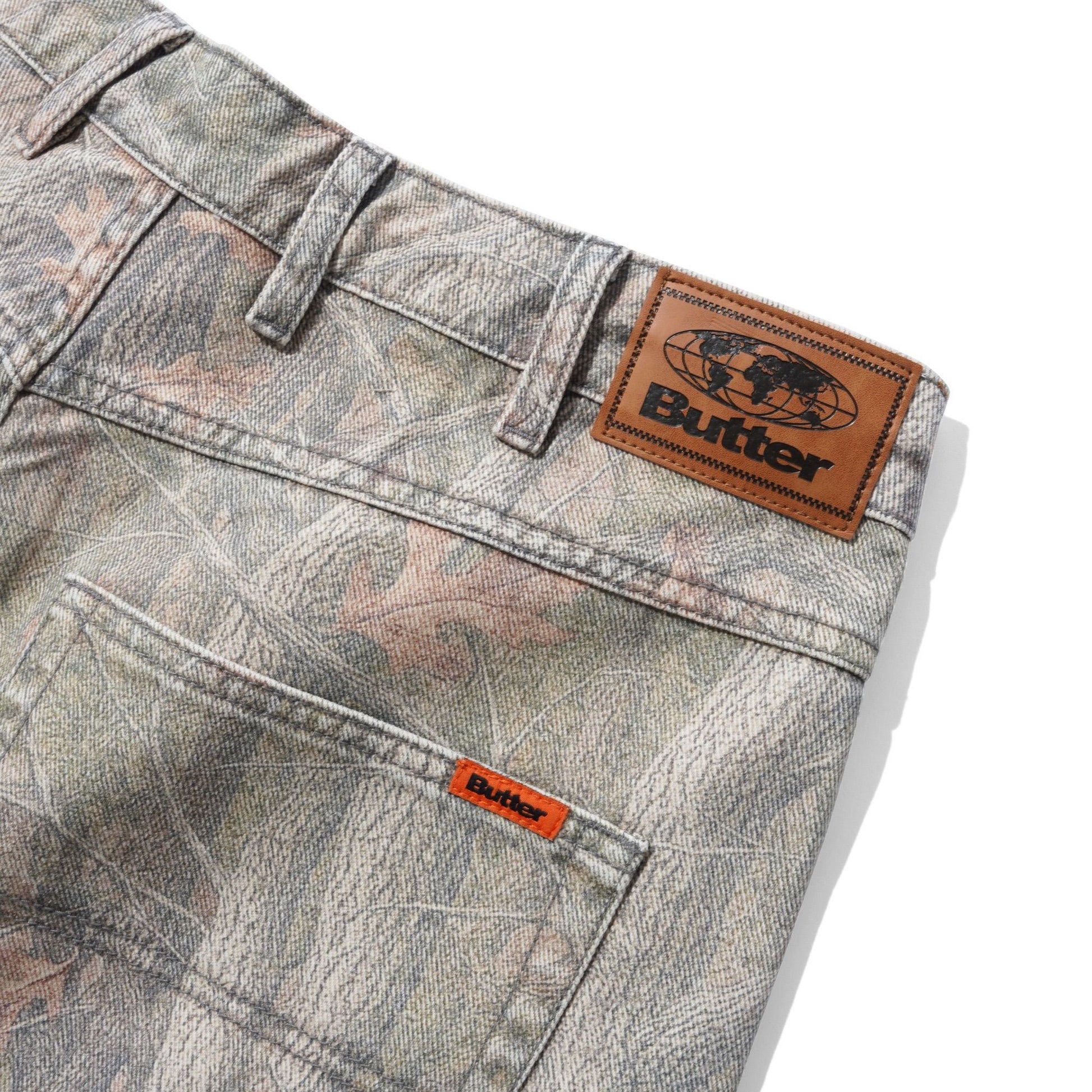 Close-up of the back waistband and pocket area of a pair of jeans in a gray and brown camouflage pattern, featuring a brown leather Butter brand patch on the waistband and a small orange Butter tab on the pocket.