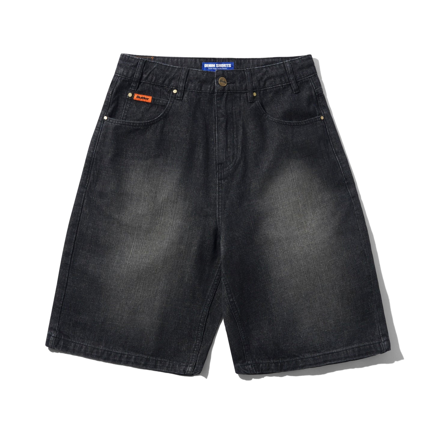 A pair of faded black denim shorts with a subtle wash, featuring classic five-pocket styling, brass-tone rivets, and a small orange brand patch on the front pocket. The brand 'Butter' is visible on the waistband label and the orange patch.