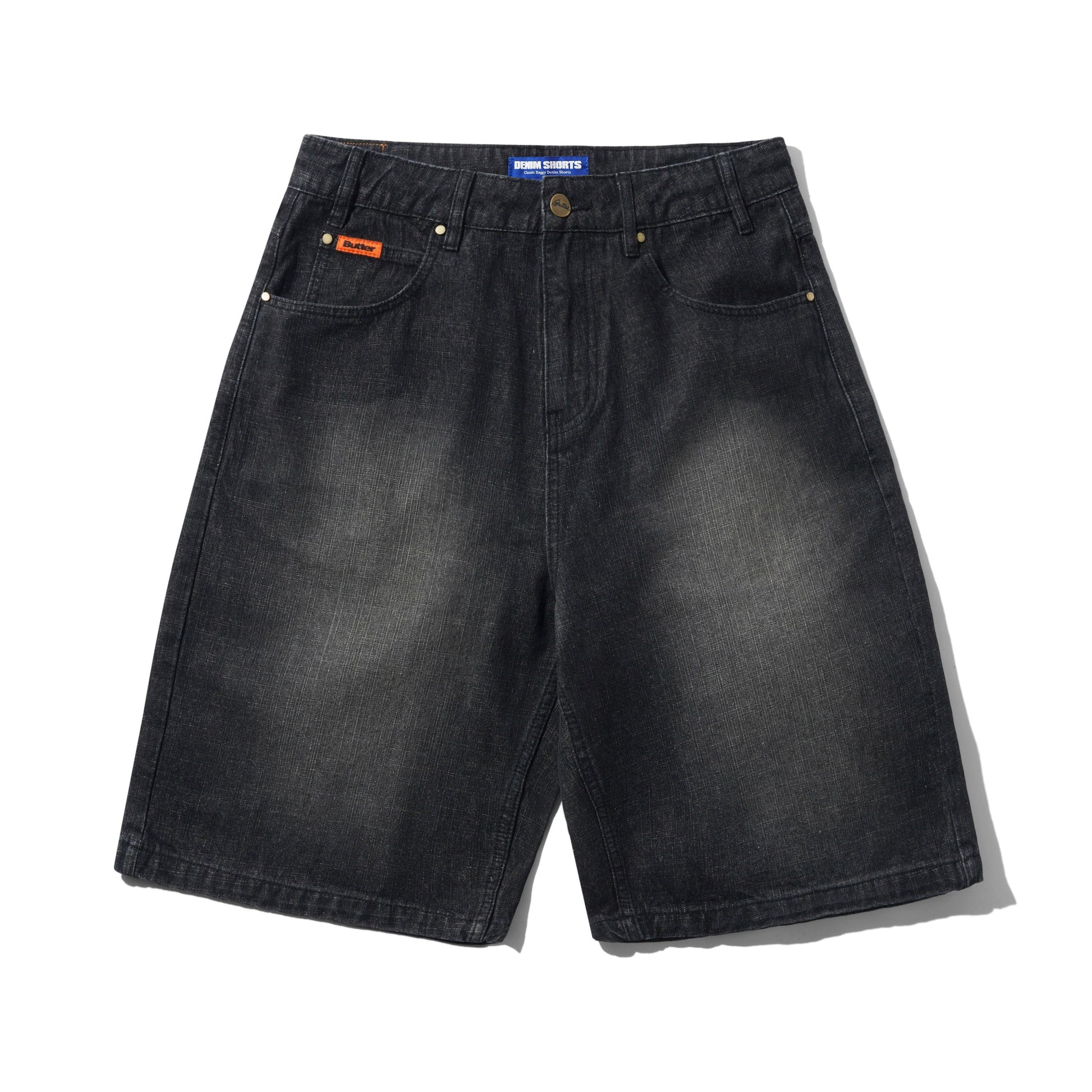 A pair of faded black denim shorts with a subtle wash, featuring classic five-pocket styling, brass-tone rivets, and a small orange brand patch on the front pocket. The brand 'Butter' is visible on the waistband label and the orange patch.