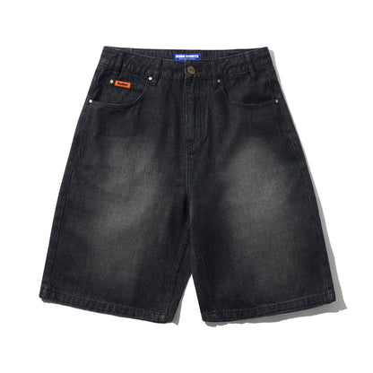 A pair of faded black denim shorts with a subtle wash, featuring classic five-pocket styling, brass-tone rivets, and a small orange brand patch on the front pocket. The brand 'Butter' is visible on the waistband label and the orange patch.