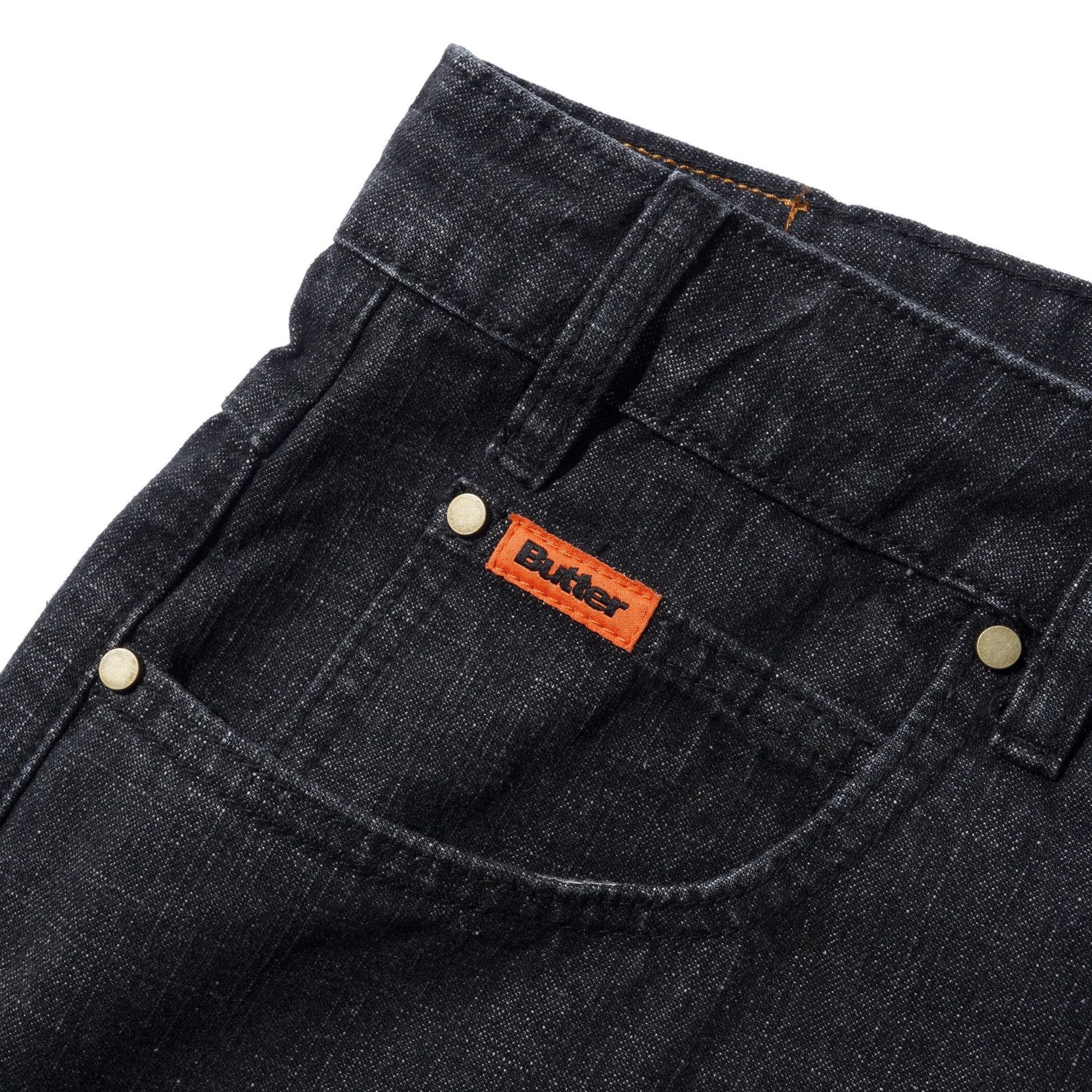 Close-up of black denim shorts showing the back pocket with a visible orange Butter brand tab and brass rivets on a white background.