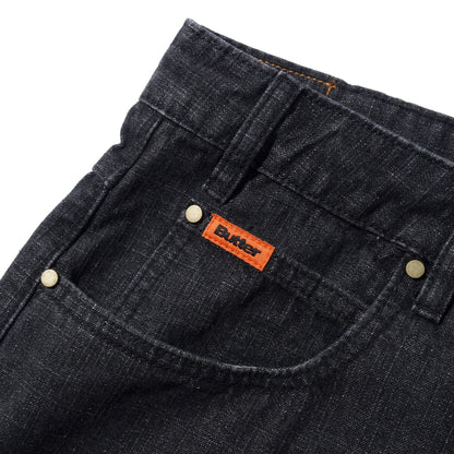 Close-up of black denim shorts showing the back pocket with a visible orange Butter brand tab and brass rivets on a white background.