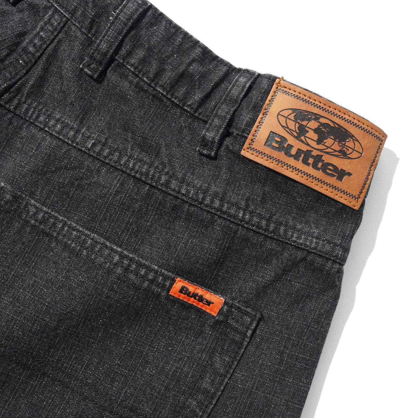 Close-up of dark gray denim jeans showing the back waistband with a large tan leather Butter patch and a smaller orange leather patch on the pocket.