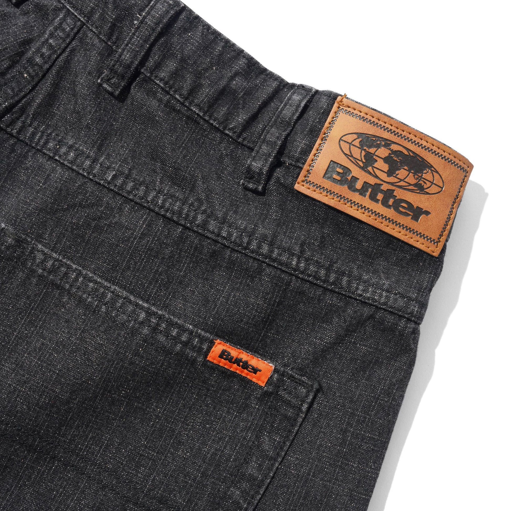 Close-up of dark gray denim jeans showing the back waistband with a large tan leather Butter patch and a smaller orange leather patch on the pocket.