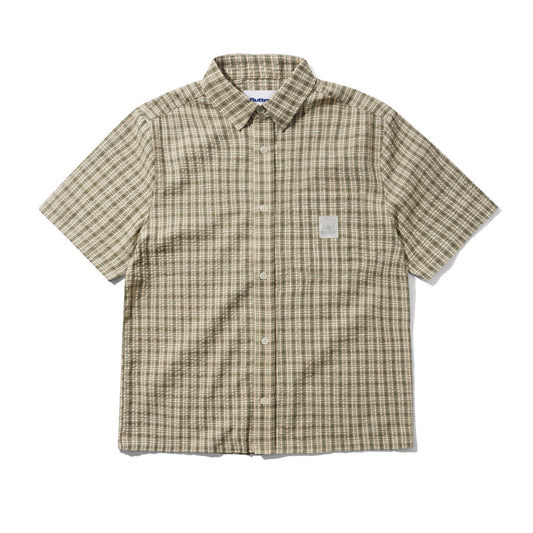 A short-sleeve button-up shirt with a beige and brown plaid pattern, featuring a small white rectangular patch on the left chest, laid flat against a white background.