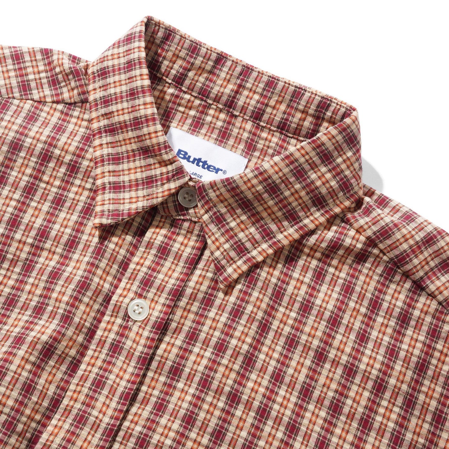 Close-up of a Butter brand plaid button-down shirt in a red, beige, and brown pattern, showing the collar, button, and inner neck label on a white background.