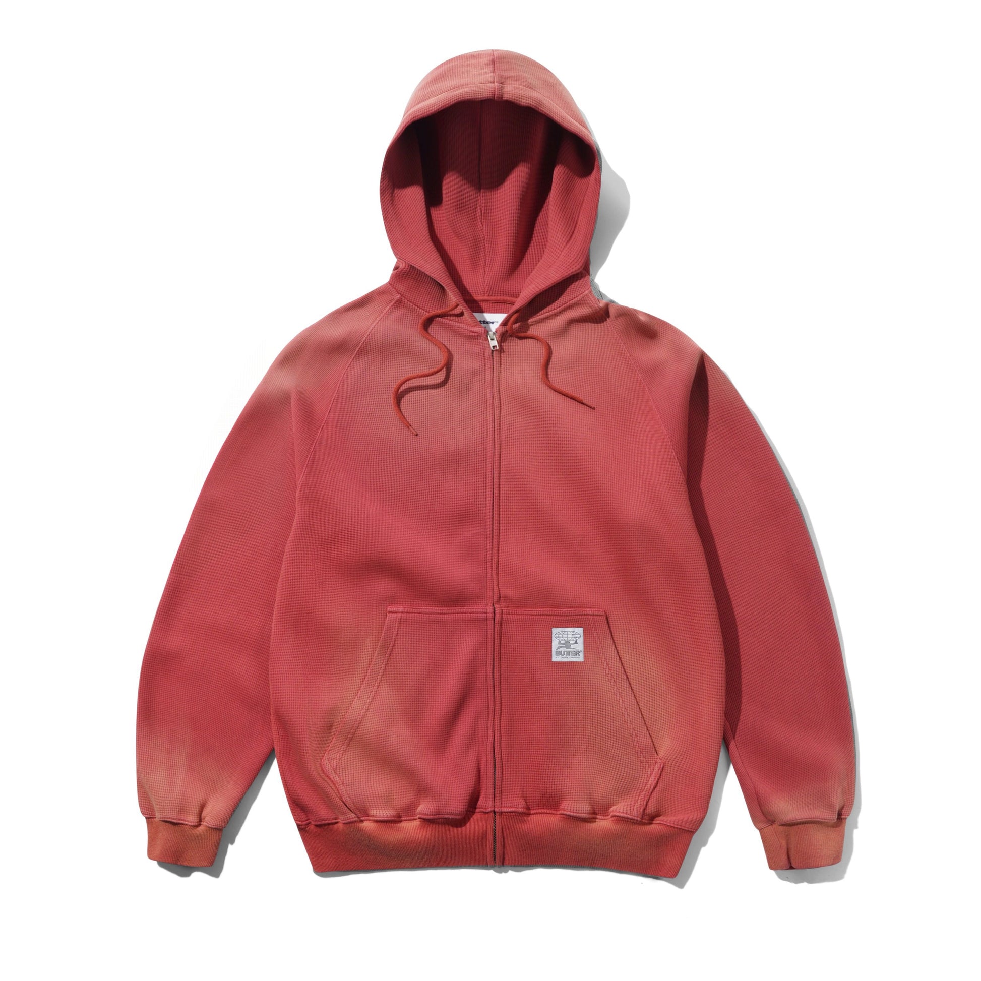 A faded red full-zip hoodie with drawstrings, a front kangaroo pocket, and a small square brand patch on the lower pocket, shown flat against a white background.