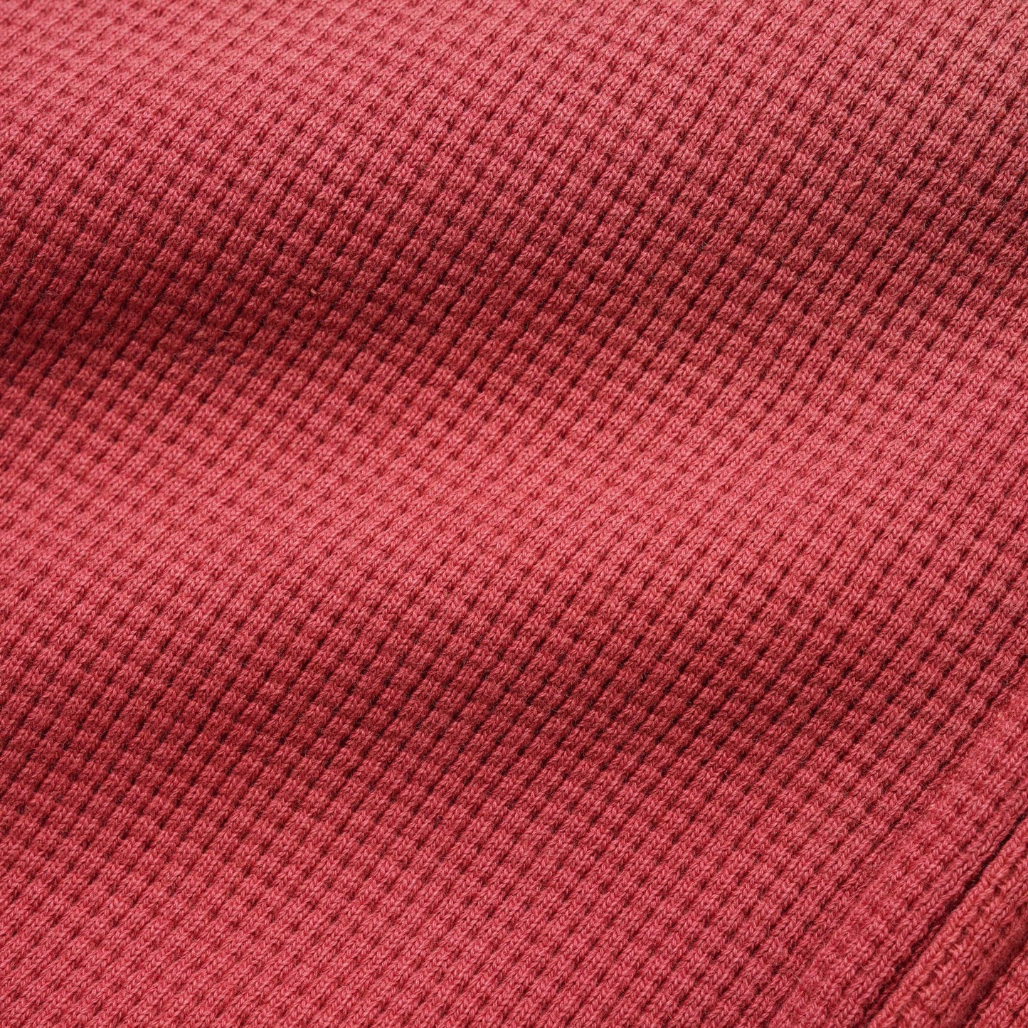A close-up of a textured waffle-knit fabric in a muted brick red or dusty rose color, showing the detailed ribbed pattern and soft folds.