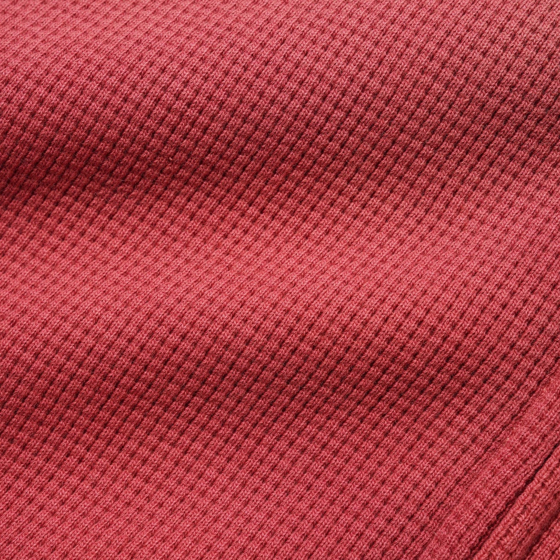 A close-up of a textured waffle-knit fabric in a muted brick red or dusty rose color, showing the detailed ribbed pattern and soft folds.