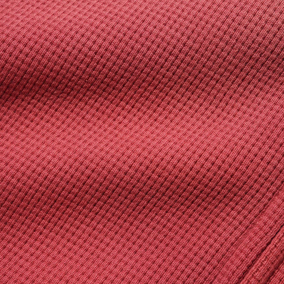 A close-up of a textured waffle-knit fabric in a muted brick red or dusty rose color, showing the detailed ribbed pattern and soft folds.