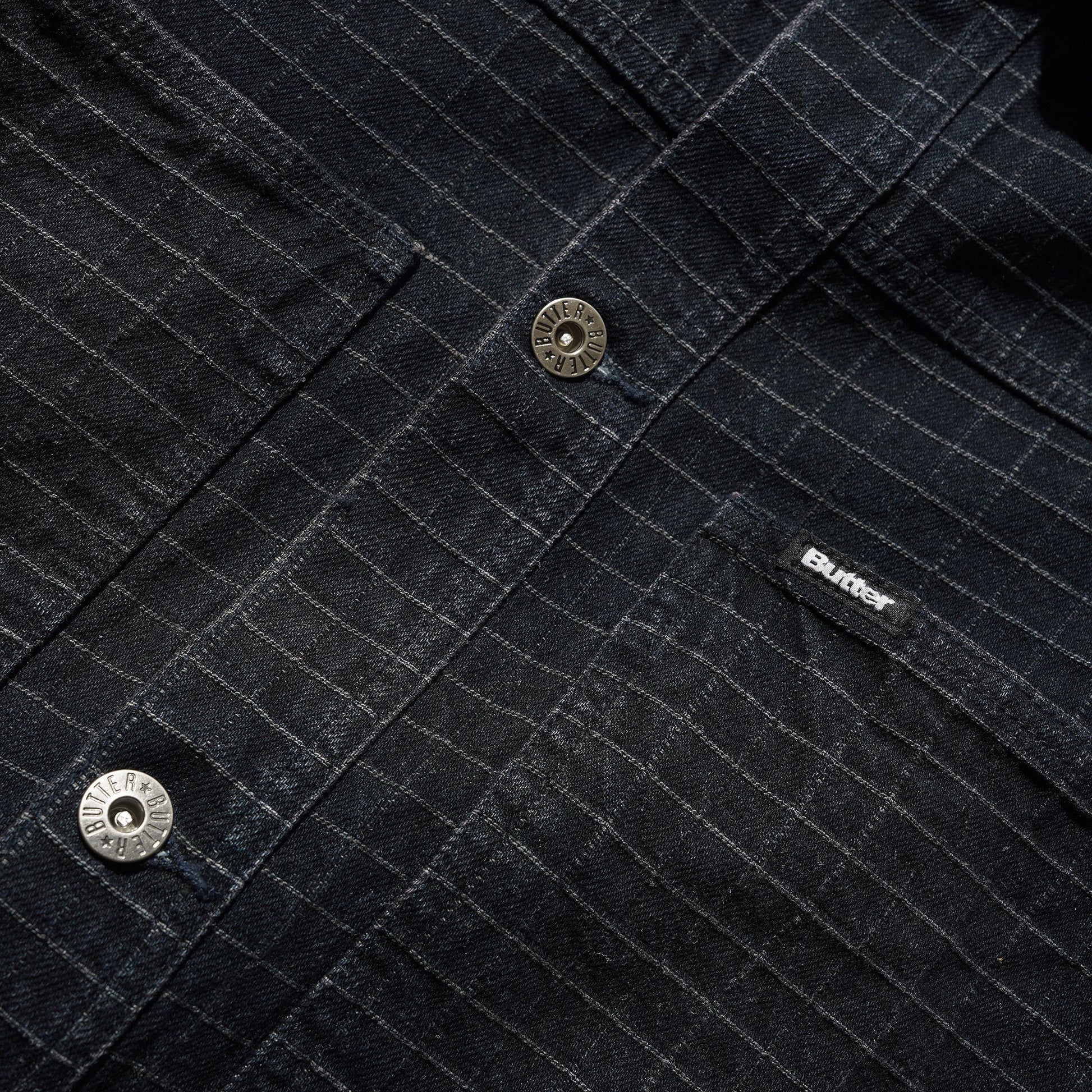 Close-up of a dark charcoal denim shirt jacket with a fine grid texture, featuring a silver-tone metal snap button and a black embroidered 'BUTTER' brand label on the chest pocket.