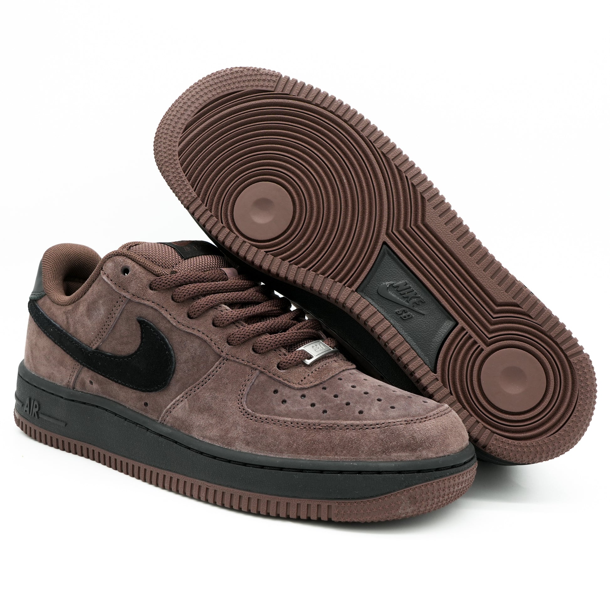 A pair of Nike Air Force 1 Low sneakers in a brown suede and black leather combination, with one shoe showing the side profile and the other showing the textured brown outsole.