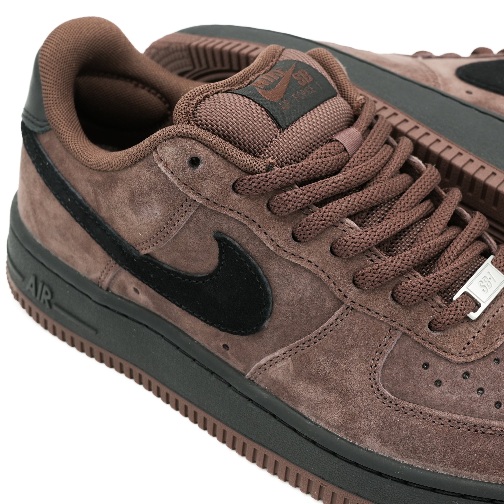 A close-up of a brown suede Nike Air Force 1 Low sneaker with a black Swoosh, matching brown laces, and a black midsole.