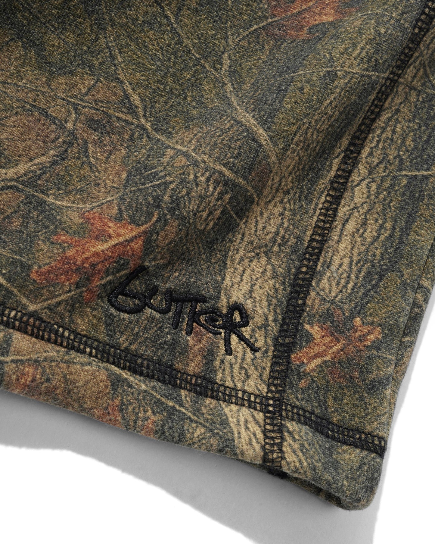 Close-up of a camouflage-patterned garment corner with black stitched edges and a black embroidered Gutker logo on a white background.