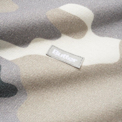 Close-up of a textured fabric with a muted camouflage pattern in beige, gray, and off-white, featuring a small rectangular stitched label that reads Butter.