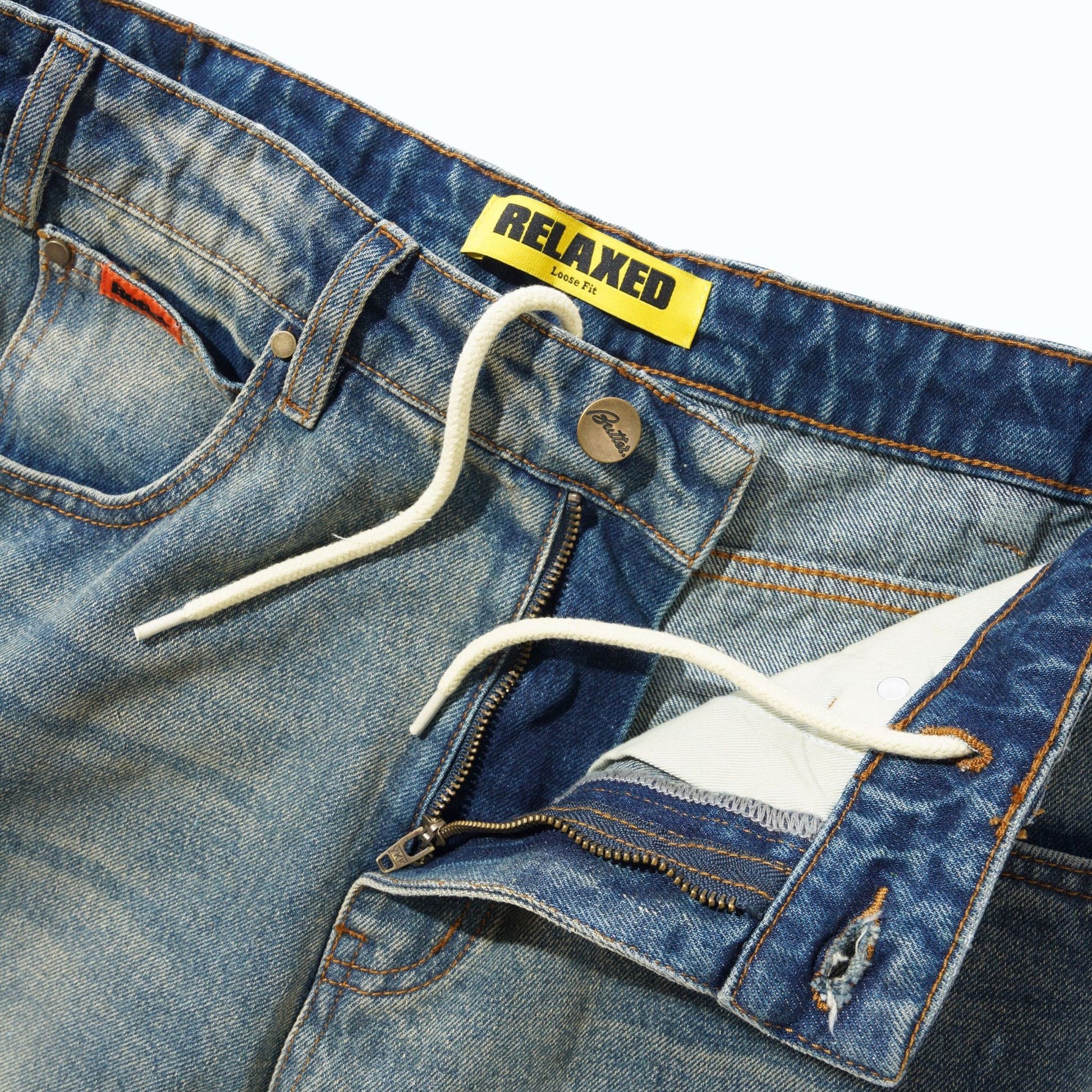 Close-up of a pair of blue denim jeans showing the waistband, white drawstring, zipper pocket detail, and a yellow RELAXED brand label.