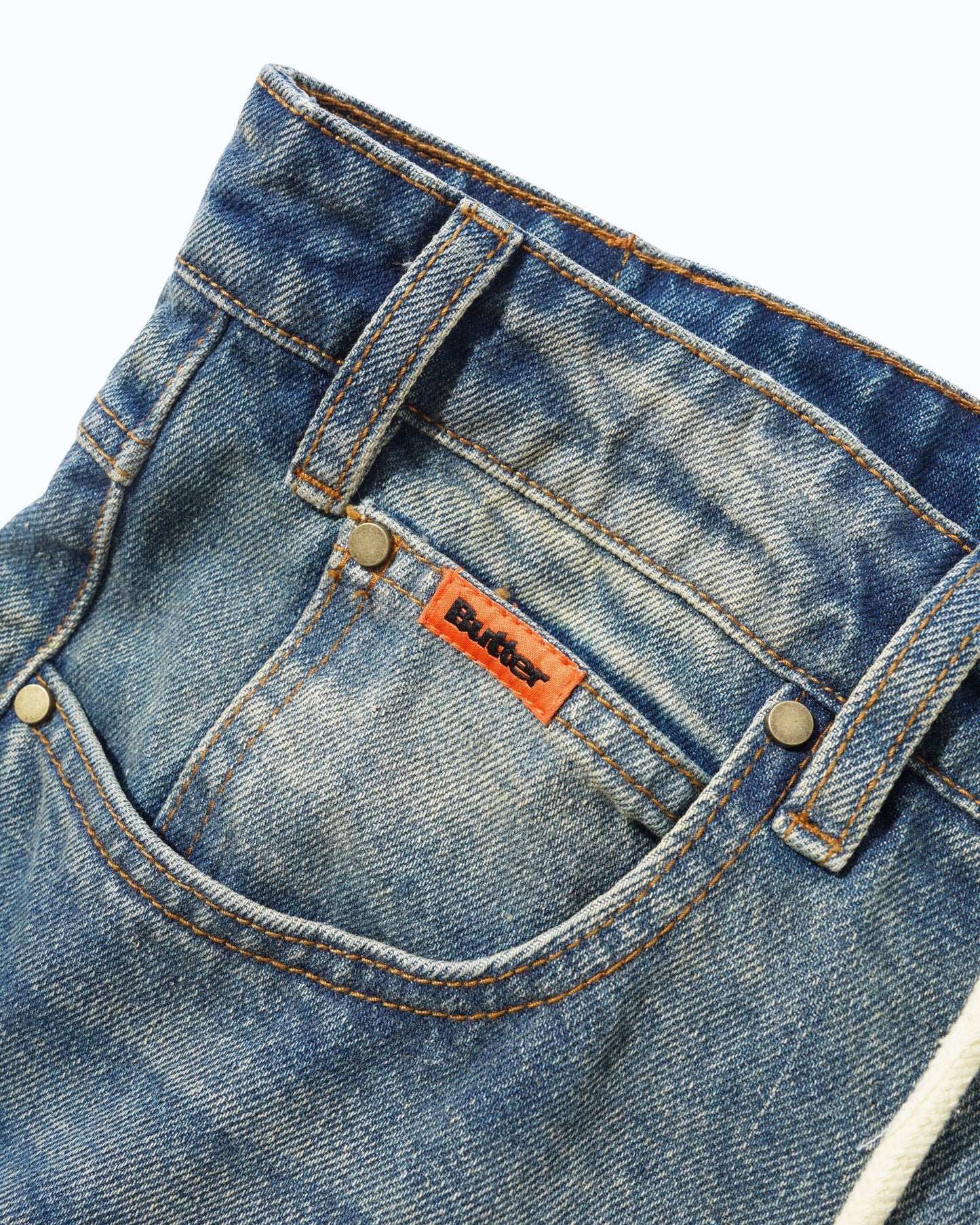 Close-up of the front pocket of blue denim jeans with a visible orange Butter brand label and metal rivets, photographed against a clean white background.