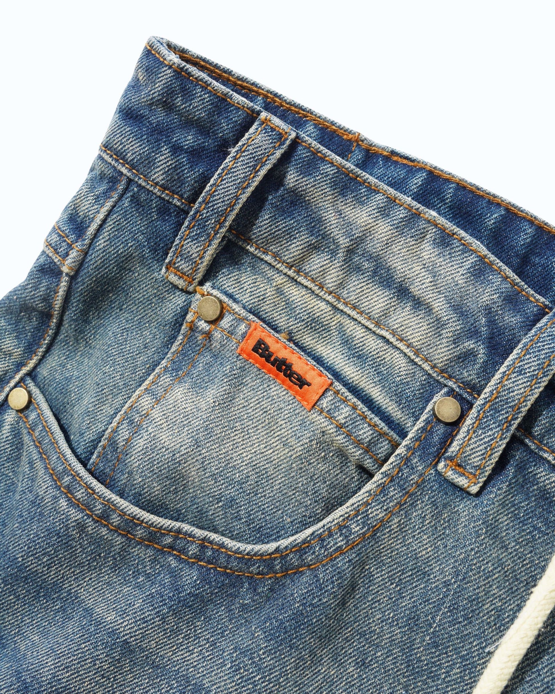 Close-up of the front pocket of blue denim jeans with a visible orange Butter brand label and metal rivets, photographed against a clean white background.