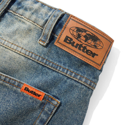 Close-up of blue denim jeans showing the back waistband, belt loops, and pocket with visible Butter brand patches on the leather and orange fabric.