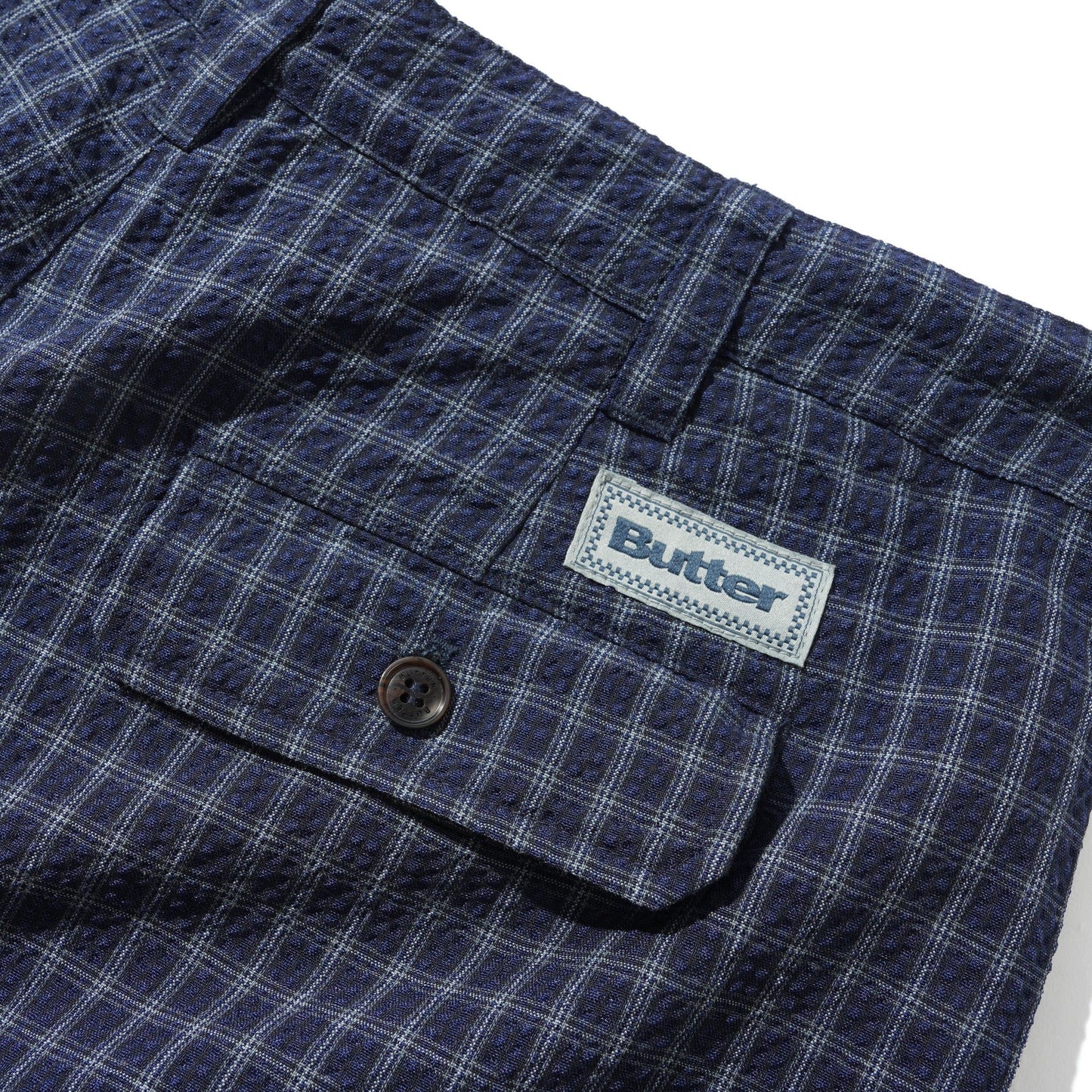 Close-up of a pair of plaid shorts in navy blue and white, featuring a buttoned pocket and a visible Butter brand label.