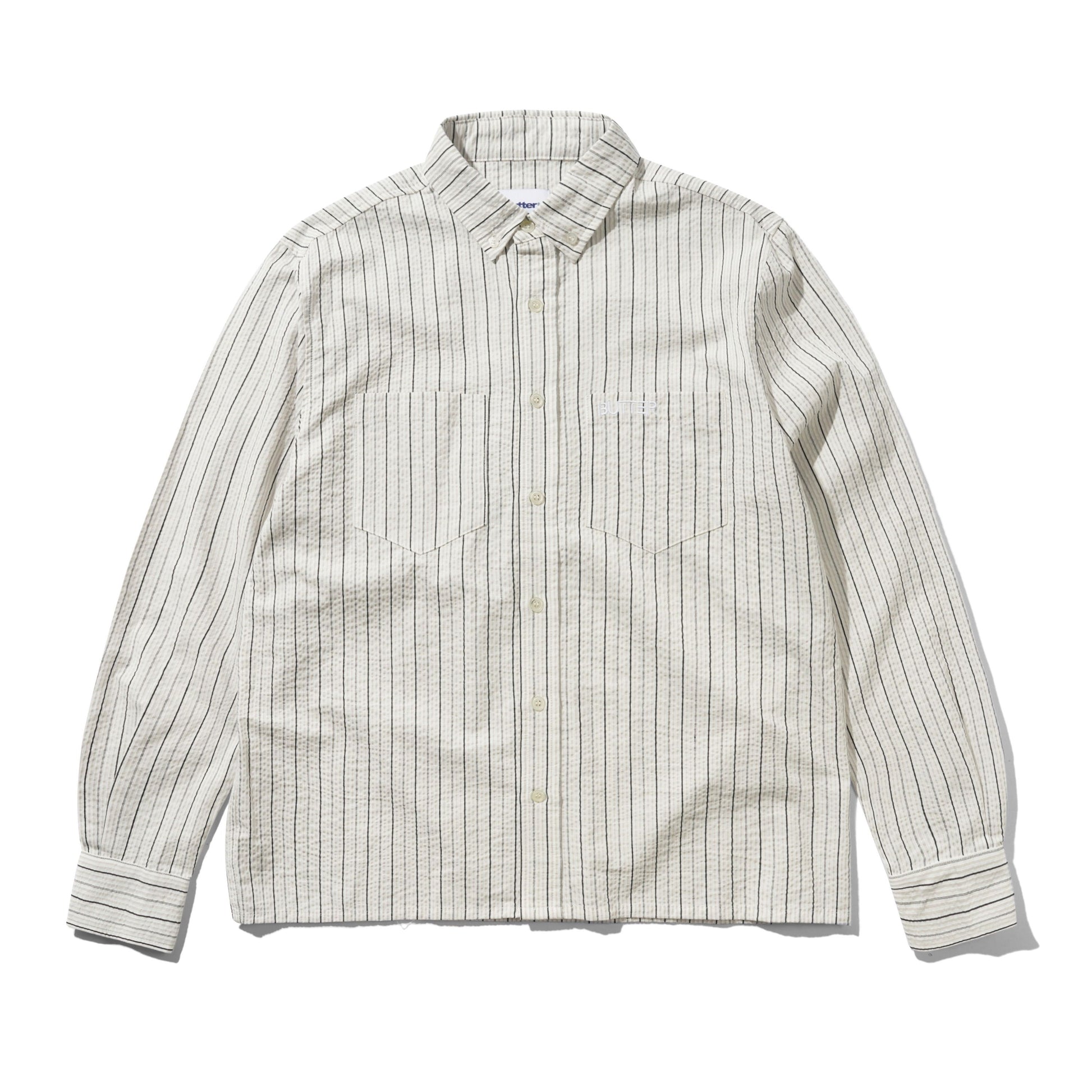 A long-sleeve button-down shirt with a textured, slightly wavy fabric, featuring vertical thin stripes in dark brown and beige on an off-white base. It has a classic collar, button-down front, and two chest pockets.