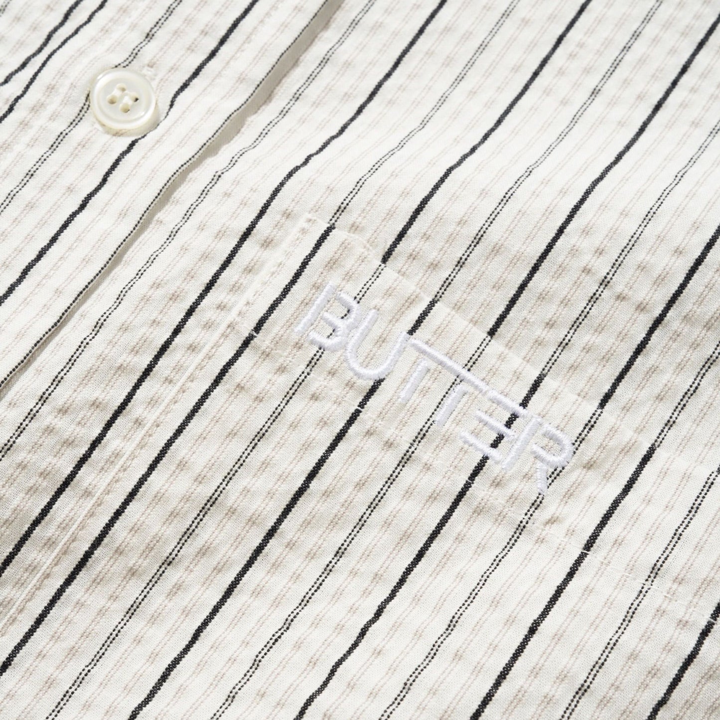 Close-up of a textured cotton shirt fabric with thin black vertical pinstripes on an off-white background, featuring a white button and a white embroidered logo that reads 'BUTTER'.
