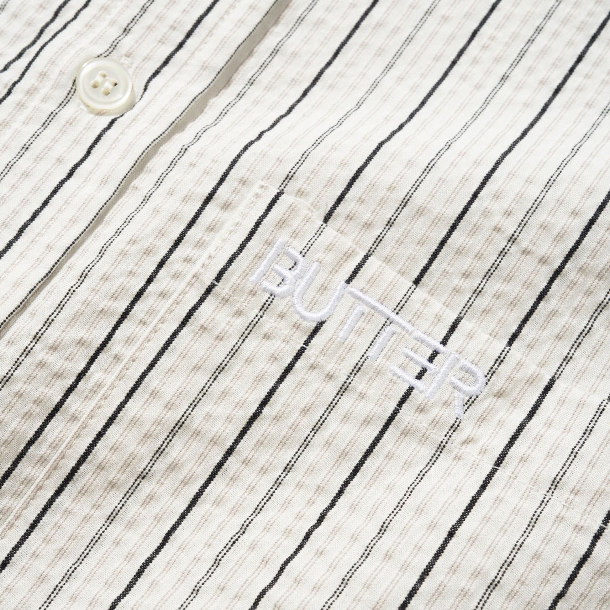 Close-up of a textured cotton shirt fabric with thin black vertical pinstripes on an off-white background, featuring a white button and a white embroidered logo that reads 'BUTTER'.