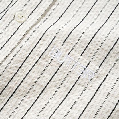 Close-up of a textured cotton shirt fabric with thin black vertical pinstripes on an off-white background, featuring a white button and a white embroidered logo that reads 'BUTTER'.