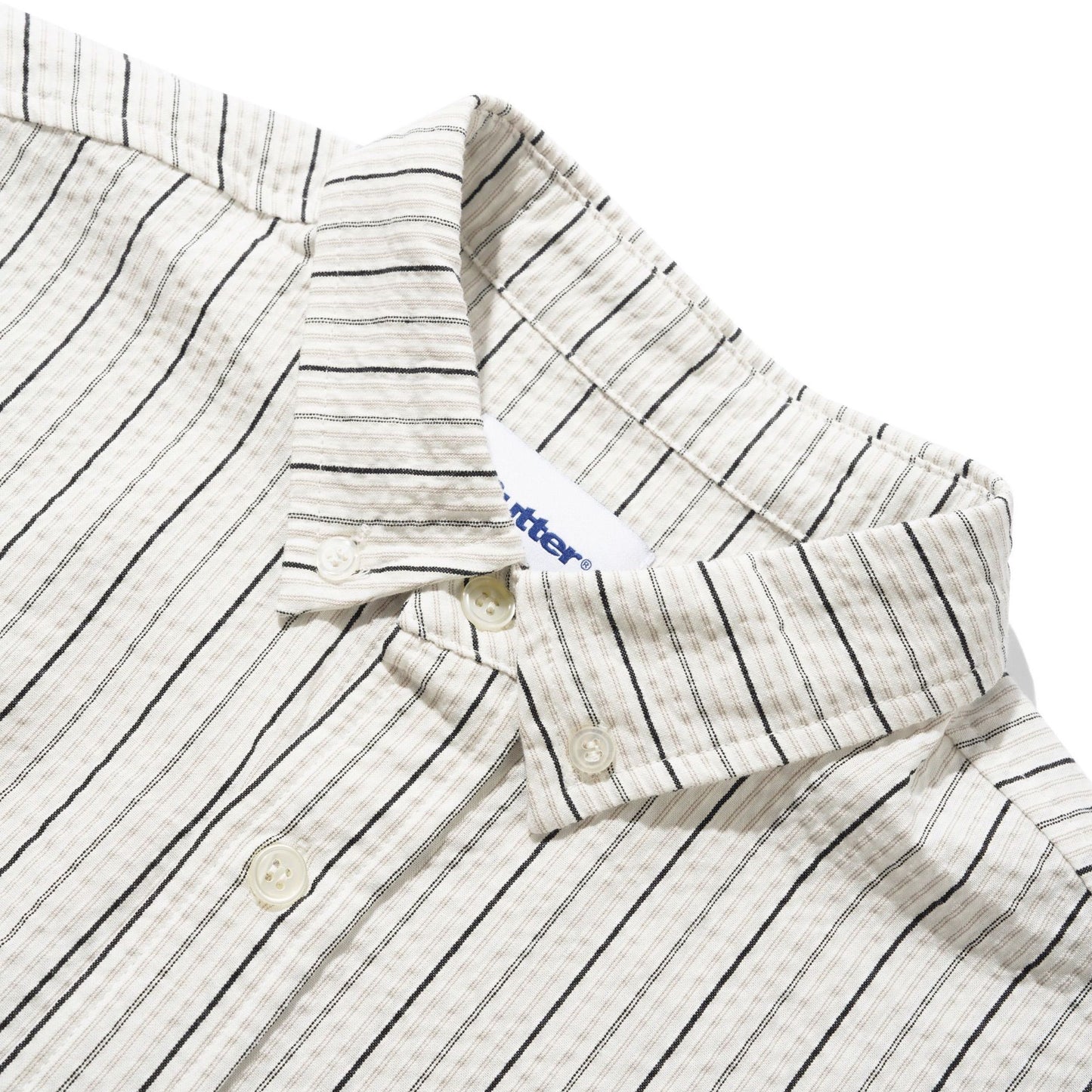 Close-up of a cotton button-down shirt collar with thin black vertical pinstripes on a cream-colored fabric, featuring a white button and a visible 'butter' brand tag.