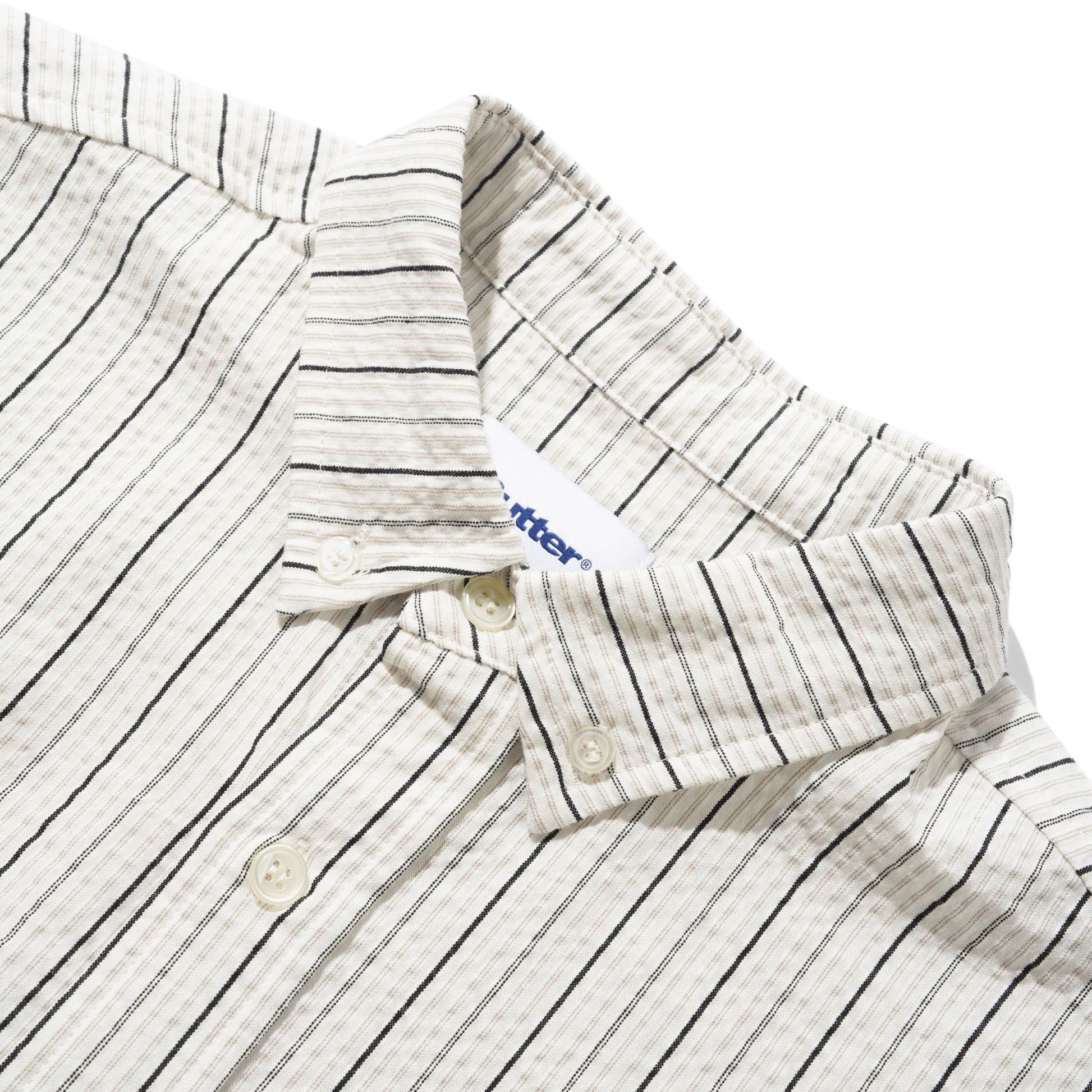 Close-up of a cotton button-down shirt collar with thin black vertical pinstripes on a cream-colored fabric, featuring a white button and a visible 'butter' brand tag.