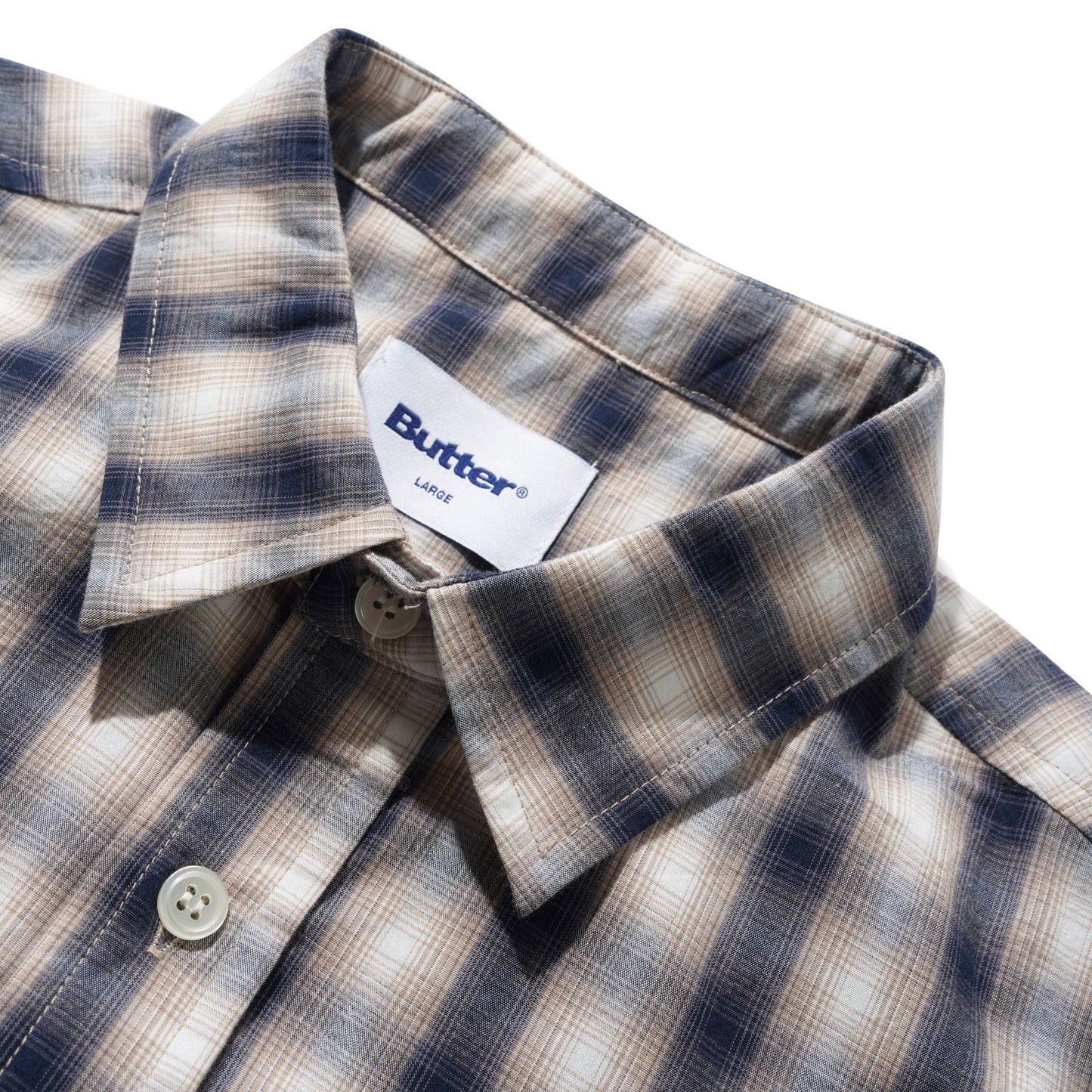 Close-up of a plaid button-down shirt collar with a visible Butter brand label. The fabric has a muted plaid pattern in beige, brown, and navy blue tones, photographed against a clean white background.