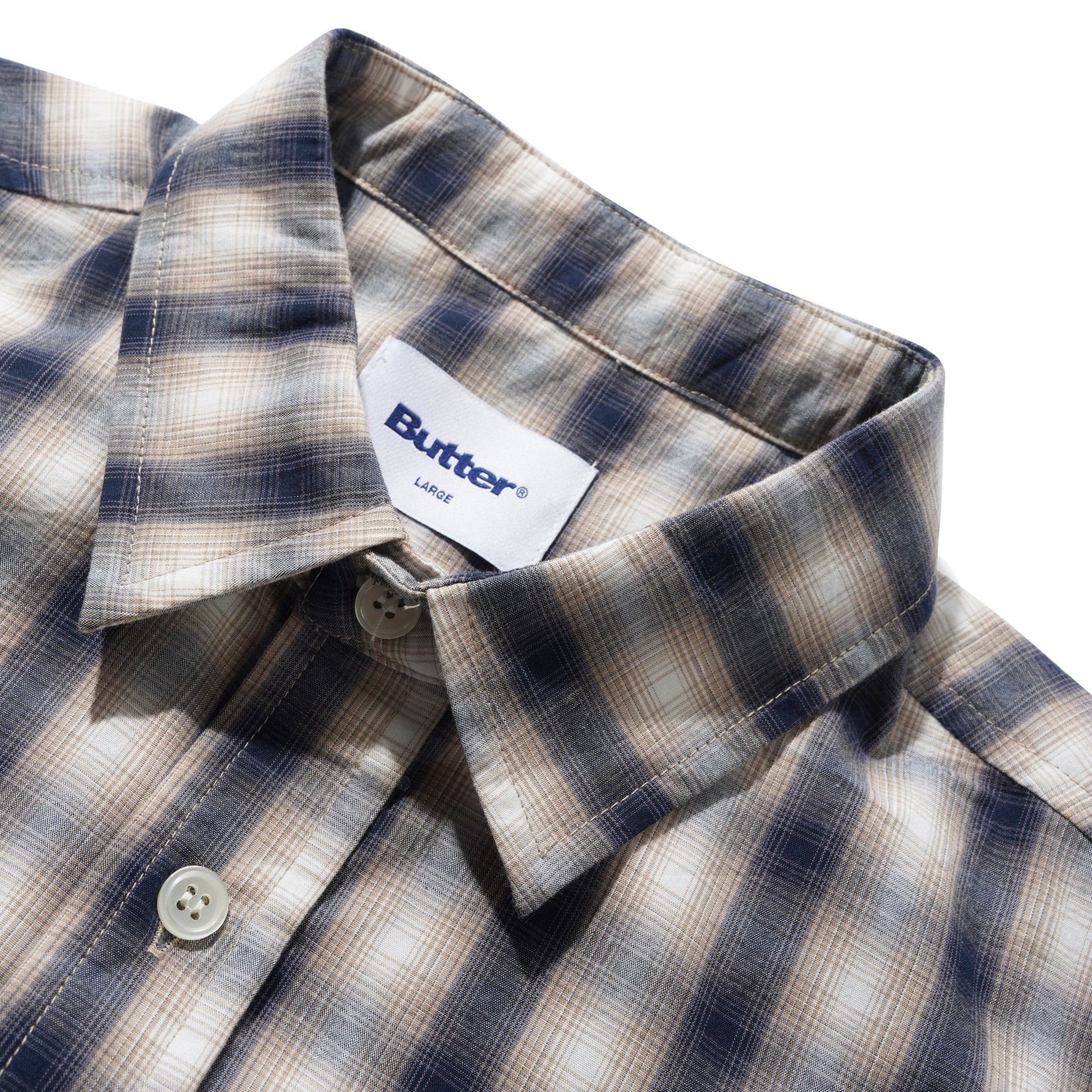 Close-up of a plaid button-down shirt collar with a visible Butter brand label. The fabric has a muted plaid pattern in beige, brown, and navy blue tones, photographed against a clean white background.