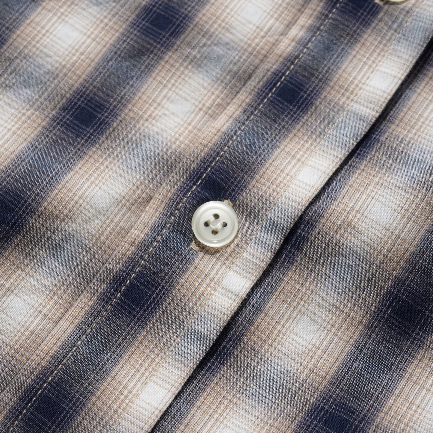Close-up of a plaid button-up shirt fabric with navy, beige, and white stripes, showing the texture and buttons.