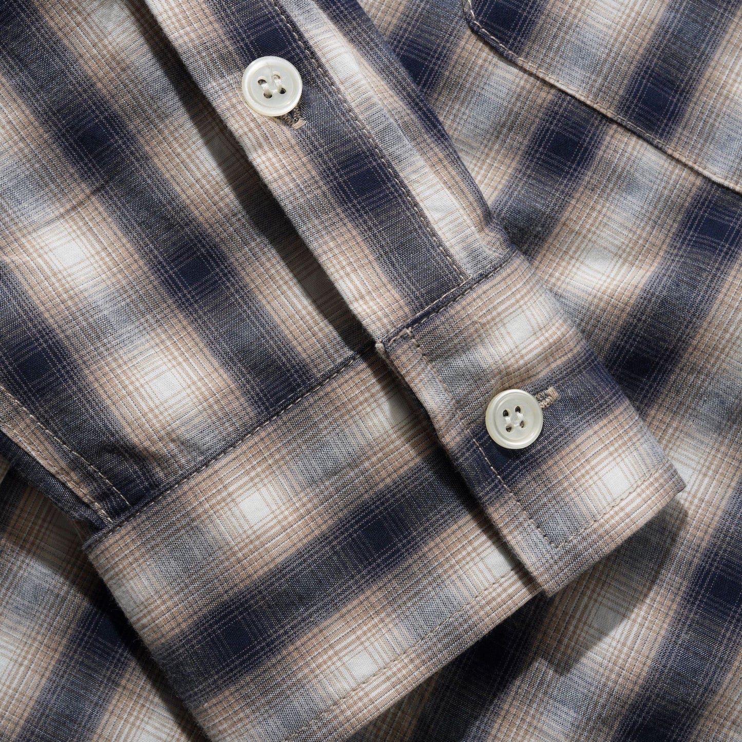 Close-up of a plaid button-up shirt showing the collar, button, cuff, and chest pocket area. The fabric has a navy, beige, and white check pattern.