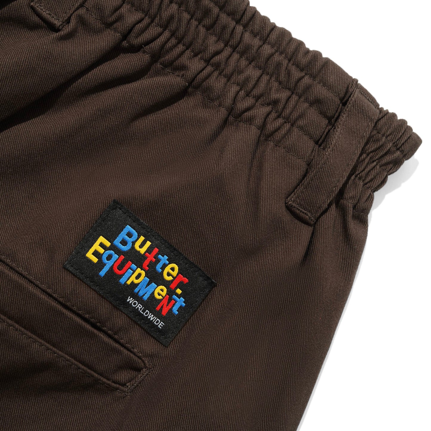 Close-up of dark brown pants showing an elastic waistband and a black embroidered patch with colorful 'Butter Equipment' text on the side pocket.