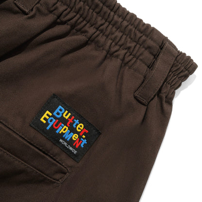Close-up of dark brown pants showing an elastic waistband and a black embroidered patch with colorful 'Butter Equipment' text on the side pocket.