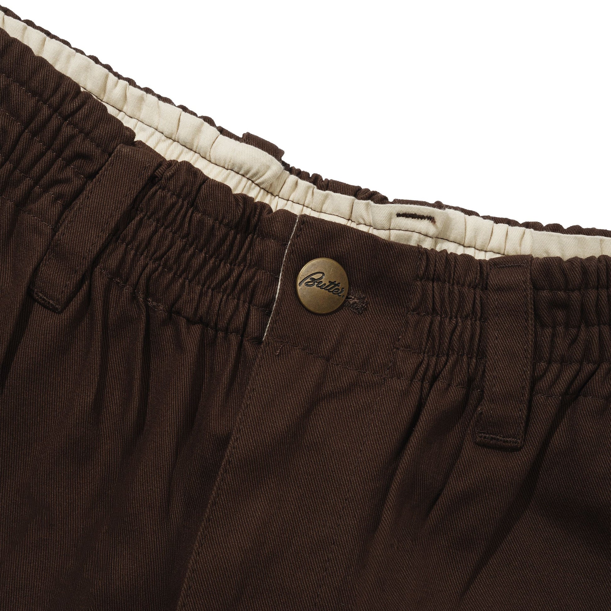 Close-up of dark brown trousers showing the elastic waistband, belt loops, a bronze button, and a cream-colored inner lining.