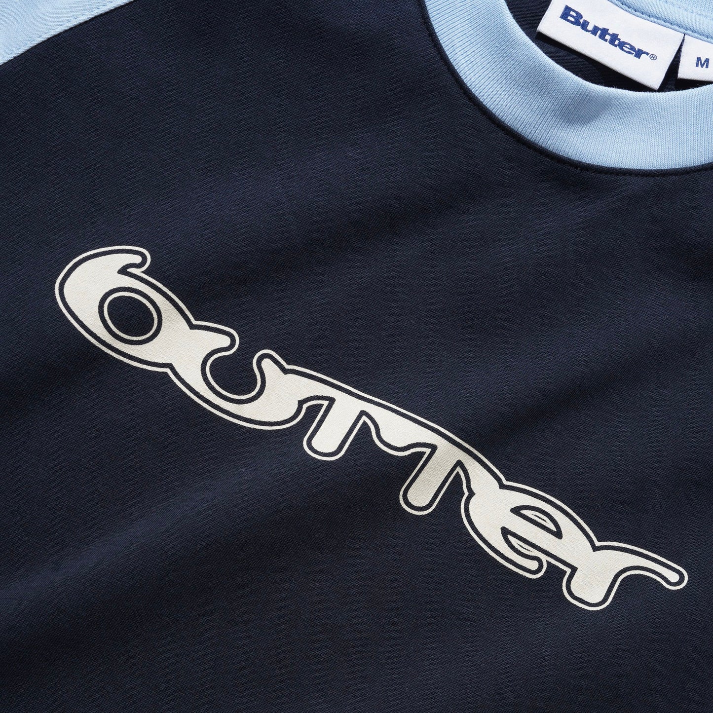 Close-up of a Butter raglan tee in navy blue with a light blue ribbed collar and a large white embroidered logo outlined in black on the chest.